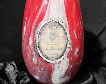 Op de afbeelding: Een beker zonder voet met een rood, wit en zwart gemarmerde afwerking. Een ovaal etiket op de voorzijde vermeldt Polyjuice Potion, From the Apothecarium of Horace E. F. Slughorn, en somt ingrediënten op zoals gaasvliegen en bloedzuigers.