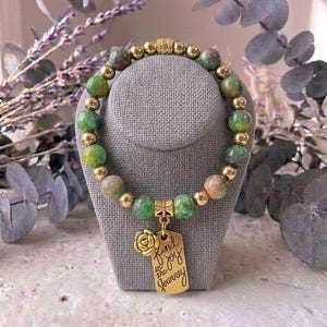 May include: A beaded bracelet featuring alternating round green and pink mottled stone beads and polished gold-tone metal spheres. A gold-tone metal charm hangs from the center, shaped like a rose and a rectangular tag inscribed with Find joy in the journey.