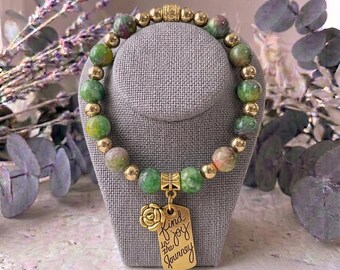 Puede incluir: Una pulsera de cuentas que alterna piedras redondas jaspeadas en verde y rosa con esferas de metal dorado pulido. Un dije de metal dorado cuelga del centro, con forma de rosa y una placa rectangular grabada con el texto Find joy in the journey.