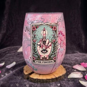 Peut inclure: Un verre à vin sans pied avec une finition marbrée rose et grise, orné d'un graphique rectangulaire central représentant une main faisant un doigt d'honneur, avec des bagues et un œil, entouré de fleurs et le texte THE FINGER.