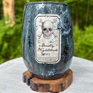 May include: A stemless tumbler with a dark blue and black marbled finish featuring blue glitter accents. A central rectangular label displays a skull and crossbones graphic, a blue rose, and the text Deadly Nightshade with additional small print.