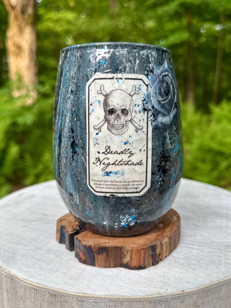 May include: A stemless tumbler with a dark blue and black marbled finish featuring blue glitter accents. A central rectangular label displays a skull and crossbones graphic, a blue rose, and the text Deadly Nightshade with additional small print.