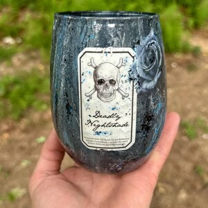 May include: A stemless tumbler with a dark, marbled blue and black finish featuring a central rectangular label. The label displays a skull and crossbones illustration above the text Deadly Nightshade and smaller printed descriptive text.