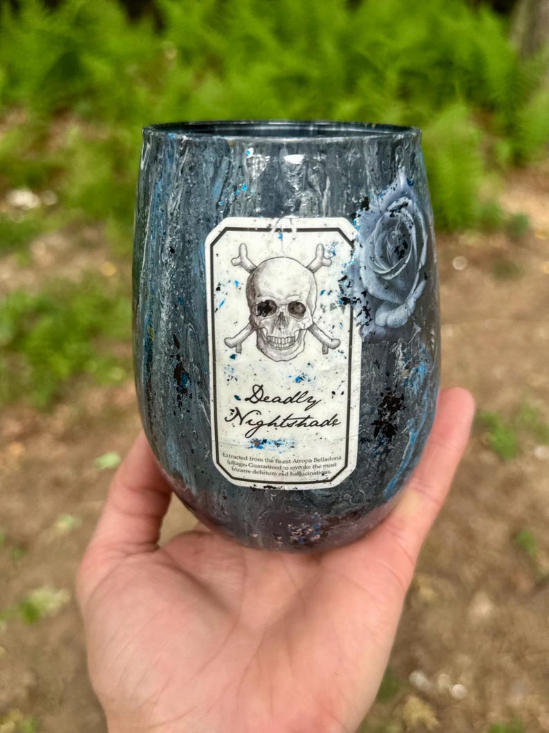 May include: A stemless tumbler with a dark, marbled blue and black finish featuring a central rectangular label. The label displays a skull and crossbones illustration above the text Deadly Nightshade and smaller printed descriptive text.