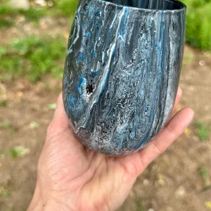 May include: A stemless tumbler featuring a marbled paint finish with swirling patterns of black, white, and metallic blue. The exterior surface has a glossy, fluid appearance with distinct color streaks and a smooth, rounded silhouette.