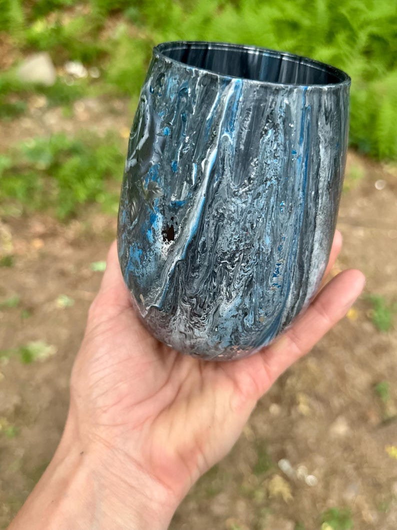 May include: A stemless tumbler featuring a marbled paint finish with swirling patterns of black, white, and metallic blue. The exterior surface has a glossy, fluid appearance with distinct color streaks and a smooth, rounded silhouette.