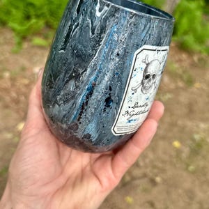 May include: A stemless tumbler with a dark blue, black, and silver marbled finish. A white rectangular label features a black ink illustration of a skull and crossbones above the text Deadly Nightshade in a cursive script font.