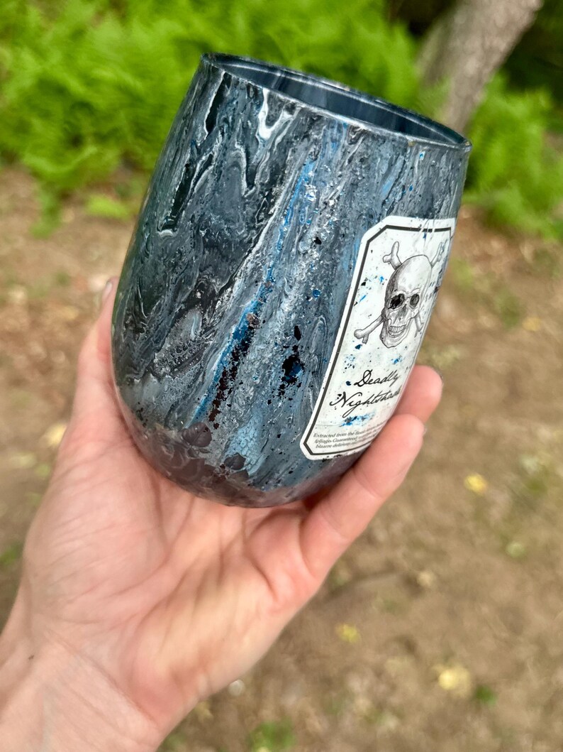 May include: A stemless tumbler with a dark blue, black, and silver marbled finish. A white rectangular label features a black ink illustration of a skull and crossbones above the text Deadly Nightshade in a cursive script font.