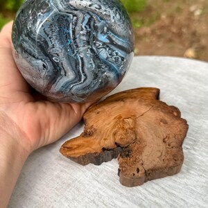 May include: A spherical object featuring a swirling marbled pattern in shades of deep blue, black, white, and grey with subtle glitter accents. Beside the sphere lies a flat, irregular piece of natural wood with visible grain and bark edges.