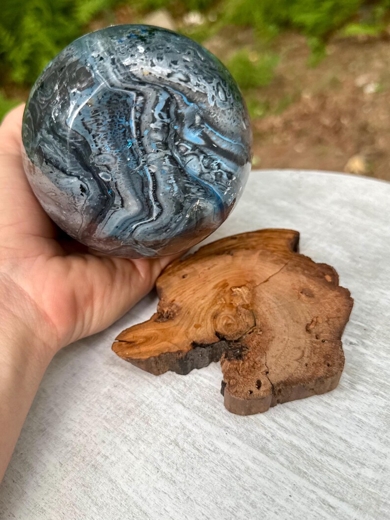 May include: A spherical object featuring a swirling marbled pattern in shades of deep blue, black, white, and grey with subtle glitter accents. Beside the sphere lies a flat, irregular piece of natural wood with visible grain and bark edges.