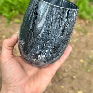 May include: A stemless tumbler featuring a fluid, marbled paint design in shades of black, white, and gray. The exterior surface displays a glossy, swirling pattern of dark and light tones that wrap around the rounded body of the drinking vessel.