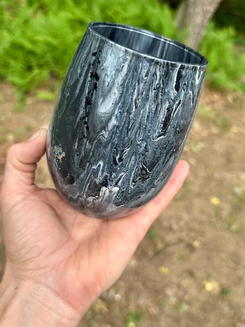 May include: A stemless tumbler featuring a fluid, marbled paint design in shades of black, white, and gray. The exterior surface displays a glossy, swirling pattern of dark and light tones that wrap around the rounded body of the drinking vessel.