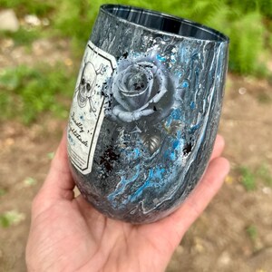 May include: A stemless tumbler featuring a dark gray and black marbled finish with vibrant blue accents. The surface displays a detailed illustration of a rose and a skull and crossbones emblem with the text Deadly Nightshade on a white label.