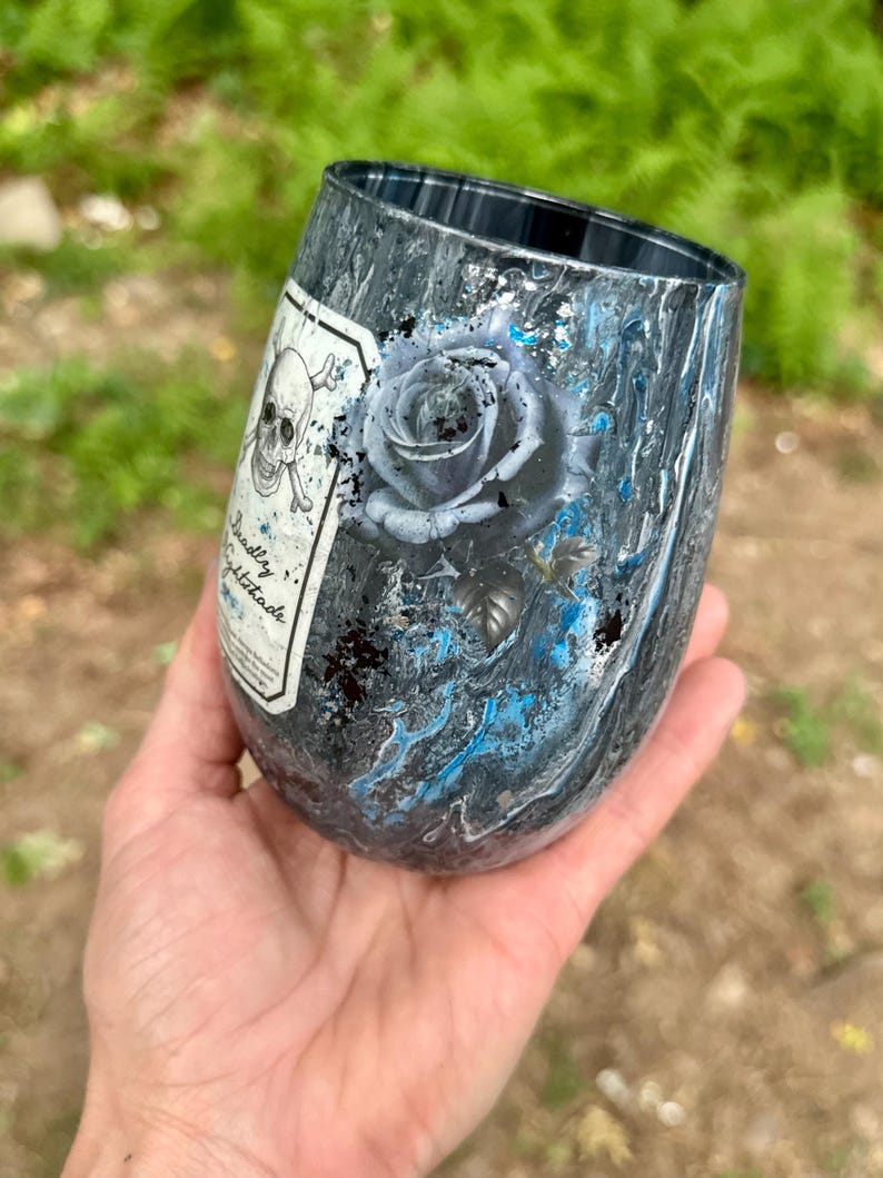 May include: A stemless tumbler featuring a dark gray and black marbled finish with vibrant blue accents. The surface displays a detailed illustration of a rose and a skull and crossbones emblem with the text Deadly Nightshade on a white label.