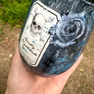 May include: A dark blue and black marbled tumbler featuring a white rectangular label with a skull and crossbones graphic and the text Deadly Nightshade. Next to the label is a detailed illustration of a blue rose with black paint splatters.