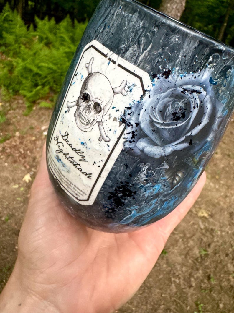 May include: A dark blue and black marbled tumbler featuring a white rectangular label with a skull and crossbones graphic and the text Deadly Nightshade. Next to the label is a detailed illustration of a blue rose with black paint splatters.