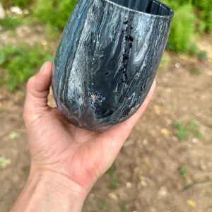 May include: A stemless tumbler featuring a marbled pattern in shades of black, gray, and white. The surface has a glossy finish with subtle flecks of blue glitter integrated into the swirling paint design, creating a fluid and dynamic appearance.