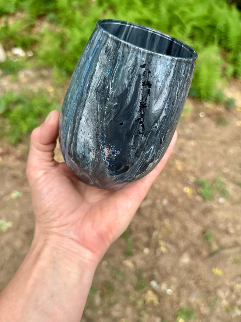 May include: A stemless tumbler featuring a marbled pattern in shades of black, gray, and white. The surface has a glossy finish with subtle flecks of blue glitter integrated into the swirling paint design, creating a fluid and dynamic appearance.