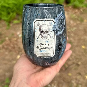 May include: A stemless tumbler with a dark, marbled finish in shades of black, grey, and blue. A rectangular label on the front features a skull and crossbones illustration above the text Deadly Nightshade, with a dark rose graphic to the side.