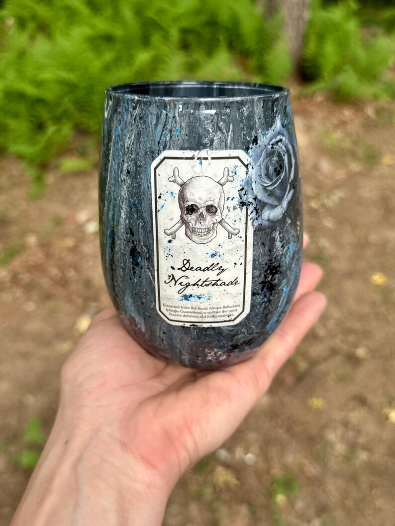 May include: A stemless tumbler with a dark, marbled finish in shades of black, grey, and blue. A rectangular label on the front features a skull and crossbones illustration above the text Deadly Nightshade, with a dark rose graphic to the side.