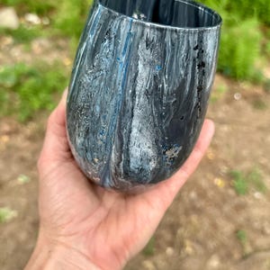 May include: A stemless tumbler featuring a marbled paint finish in shades of black, white, and metallic blue. The exterior surface displays swirling patterns of dark and light colors with a glossy texture, creating a fluid and abstract visual effect.