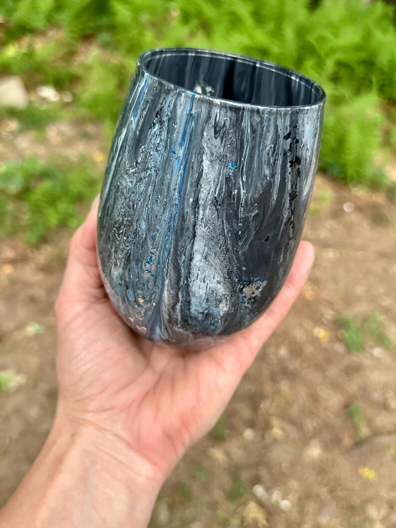 May include: A stemless tumbler featuring a marbled paint finish in shades of black, white, and metallic blue. The exterior surface displays swirling patterns of dark and light colors with a glossy texture, creating a fluid and abstract visual effect.