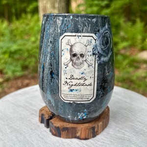 May include: A stemless tumbler with a dark, marbled finish in shades of black, grey, and blue, featuring a rectangular label with a skull and crossbones graphic and the text "Deadly Nightshade" along with smaller printed descriptive text.