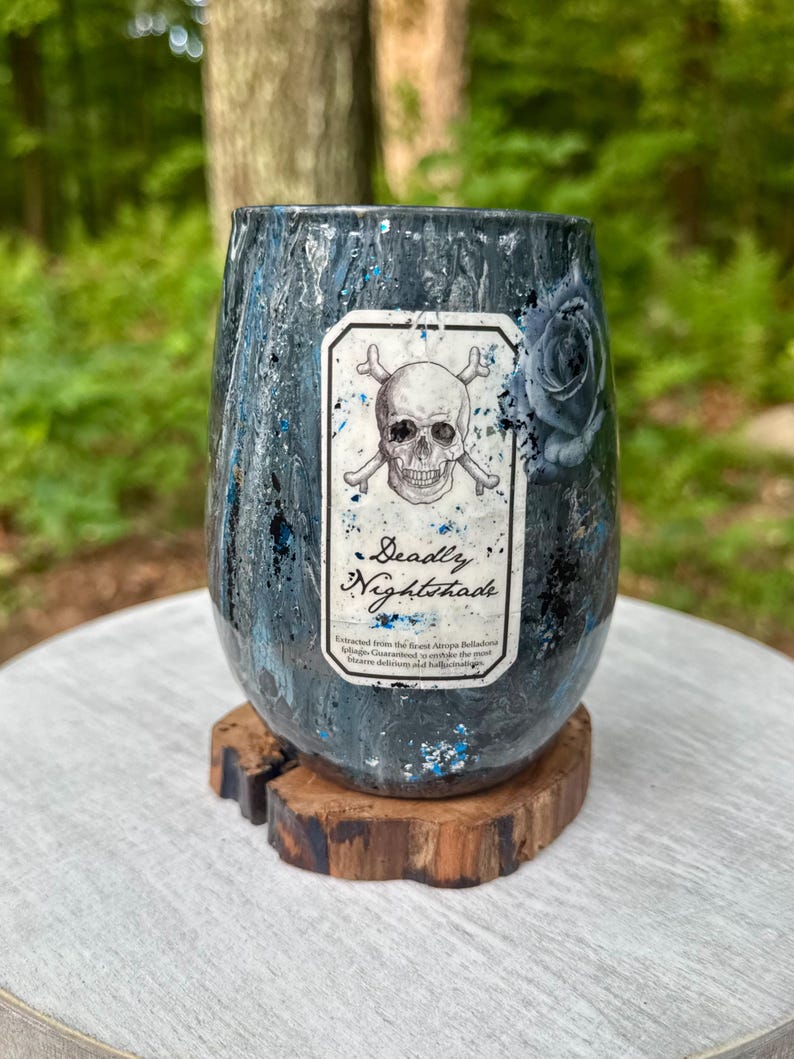 May include: A stemless tumbler with a dark, marbled finish in shades of black, grey, and blue, featuring a rectangular label with a skull and crossbones graphic and the text "Deadly Nightshade" along with smaller printed descriptive text.