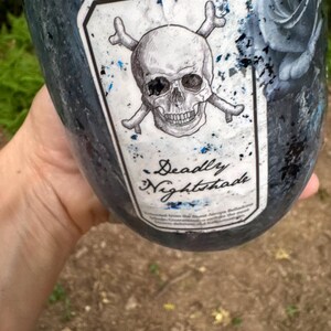 May include: A dark blue and black tumbler featuring a white rectangular label with a skull and crossbones illustration. The label includes the text Deadly Nightshade in a script font. The tumbler surface has a textured finish with blue glitter.