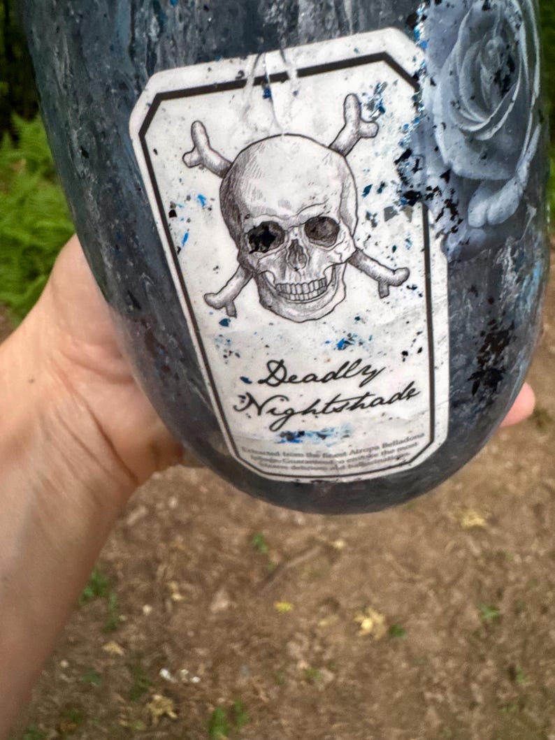May include: A dark blue and black tumbler featuring a white rectangular label with a skull and crossbones illustration. The label includes the text Deadly Nightshade in a script font. The tumbler surface has a textured finish with blue glitter.