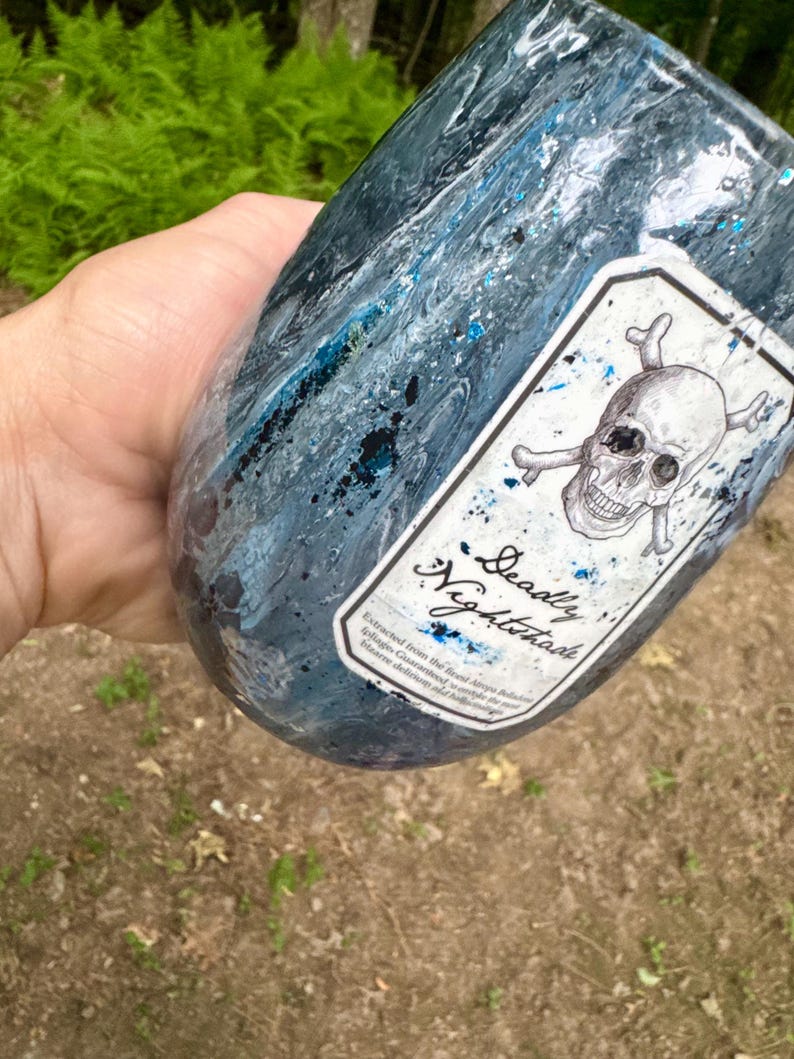 May include: A dark blue and black marbled tumbler featuring a white rectangular label with a skull and crossbones illustration. The label includes the text Deadly Nightshade and smaller printed text below. The surface has a glossy finish.