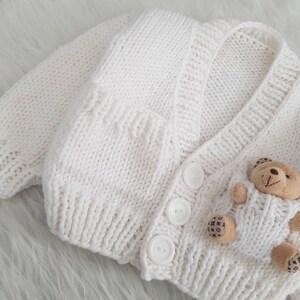 May include: White knitted baby cardigan with three white buttons and a small brown teddy bear attached to the front.