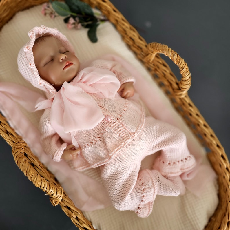 May include: A pink knitted baby outfit with a matching bonnet and a pink satin bow. The outfit is laid out in a wicker basket.