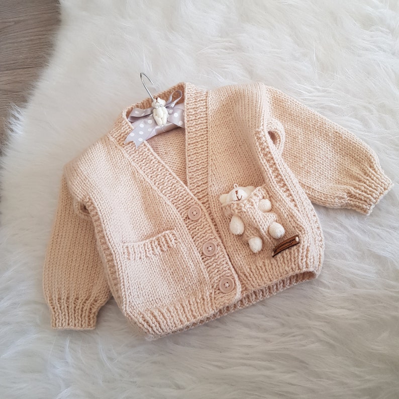 May include: A cream-colored knitted cardigan with a small teddy bear sewn onto the pocket. The cardigan has a V-neck and button closure.