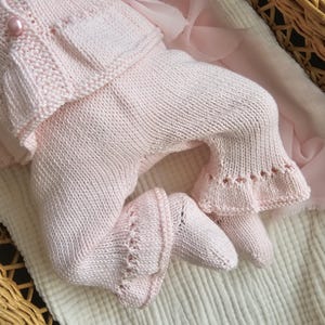 May include: A pink knitted baby outfit with a ruffled hem and a button on the front.