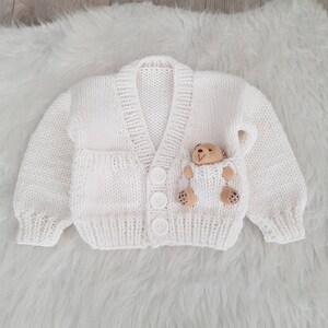 May include: White knitted baby cardigan with a button closure and a small teddy bear sewn onto the front pocket.