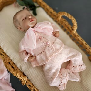 May include: A pink knitted baby outfit with a matching pink bow. The outfit is made of soft, delicate yarn and features a button-up cardigan and pants with lace detailing. The baby doll is lying in a wicker basket.