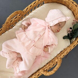 May include: A pink knitted baby outfit with a matching hat, featuring a delicate lace pattern and pearl buttons. The outfit is laid out in a wicker basket.