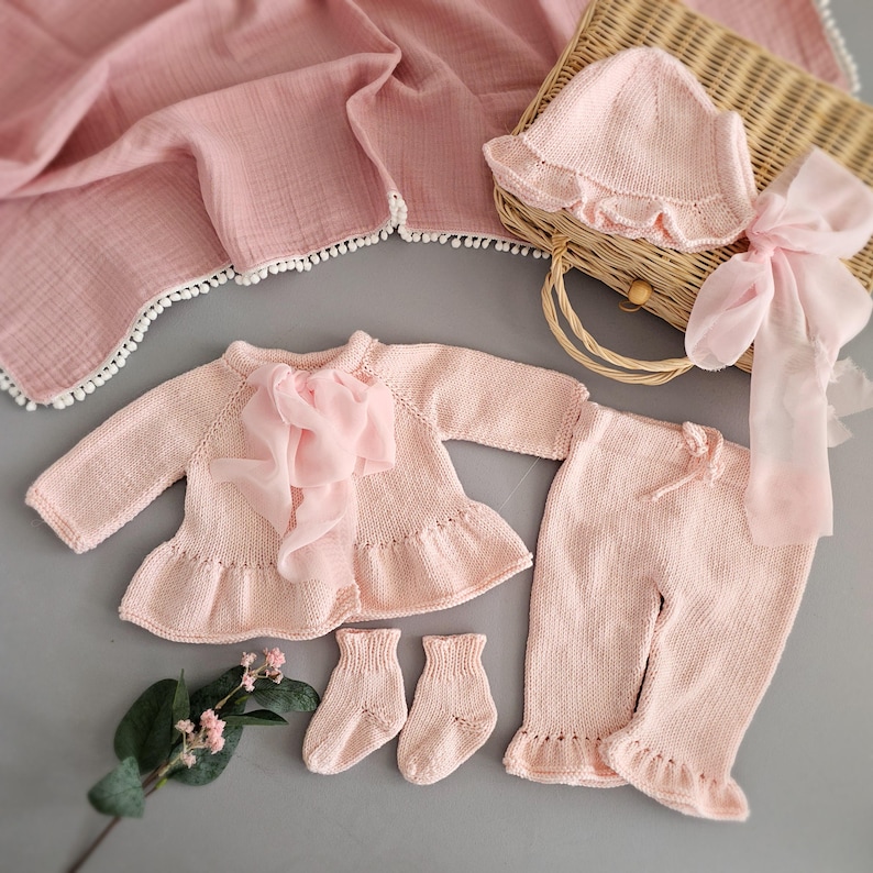 May include: A pink knitted baby outfit set with a cardigan, pants, socks, and a hat. The set is decorated with ruffles and a pink ribbon.