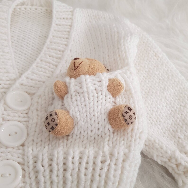 May include: White knitted cardigan with a teddy bear sewn into the pocket. The cardigan has a V-neck and button closure.