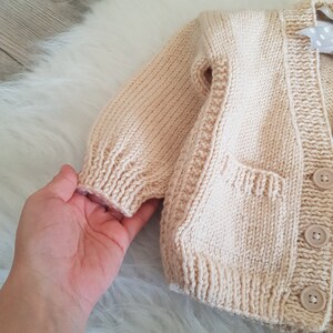 May include: A cream-colored knitted cardigan with a pocket and button closure.