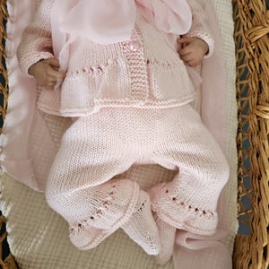 May include: A pink knitted baby outfit with a cardigan and pants. The outfit has a pearl button and delicate lace detailing.