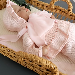 May include: A pink knitted baby outfit with a matching bonnet and a sheer pink scarf. The outfit is laid out in a wicker basket.
