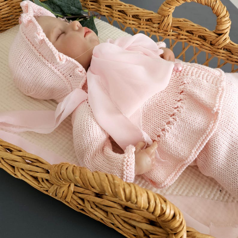 May include: A pink knitted baby outfit with a matching bonnet and a sheer pink scarf. The outfit is laid out in a wicker basket.