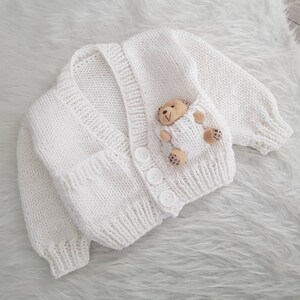 May include: White knitted baby cardigan with a small teddy bear sewn onto the front pocket.