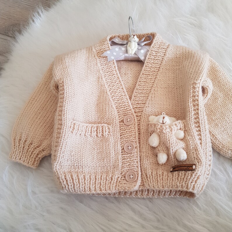 May include: A pale pink knitted cardigan with a small teddy bear sewn onto the pocket. The cardigan has a V-neck and buttons down the front.