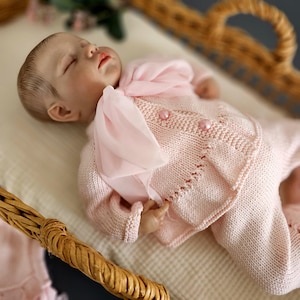 May include: A sleeping baby doll wearing a pink knitted outfit with a matching pink ribbon. The doll is lying in a wicker basket.
