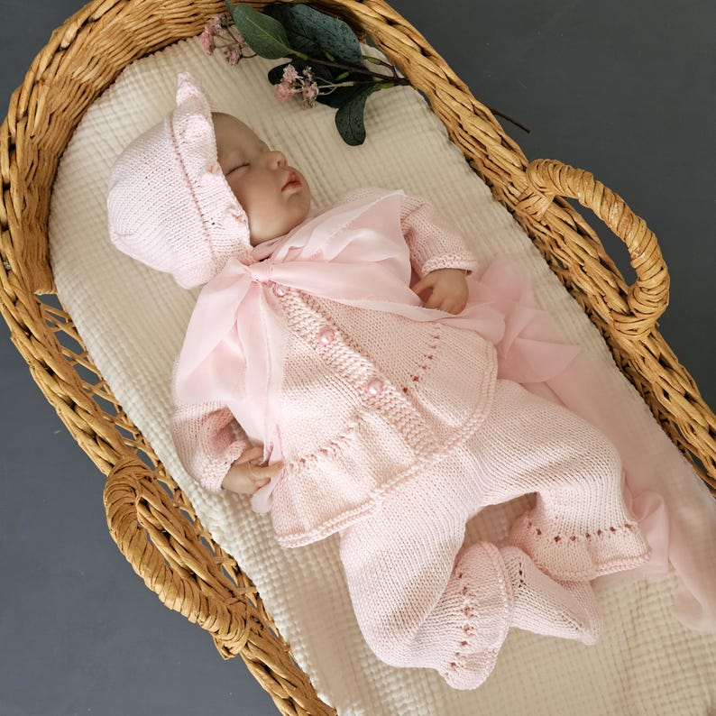 May include: A pink knitted baby outfit with a matching hat, featuring a delicate lace pattern and pearl embellishments. The outfit is laid out in a wicker basket.