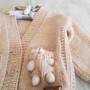 May include: A cream-colored knitted cardigan with a pocket on the front. The pocket has a small stuffed animal inside. The cardigan has buttons down the front.