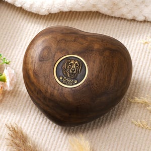 Op de afbeelding: Hartvormige houten urn met een donkerbruine afwerking. Een cirkelvormig bronzen embleem toont een hondenportret, de naam "Bruno" en de jaren "2012-2025". De urn is ontworpen voor huisdierherdenkingsdoeleinden.